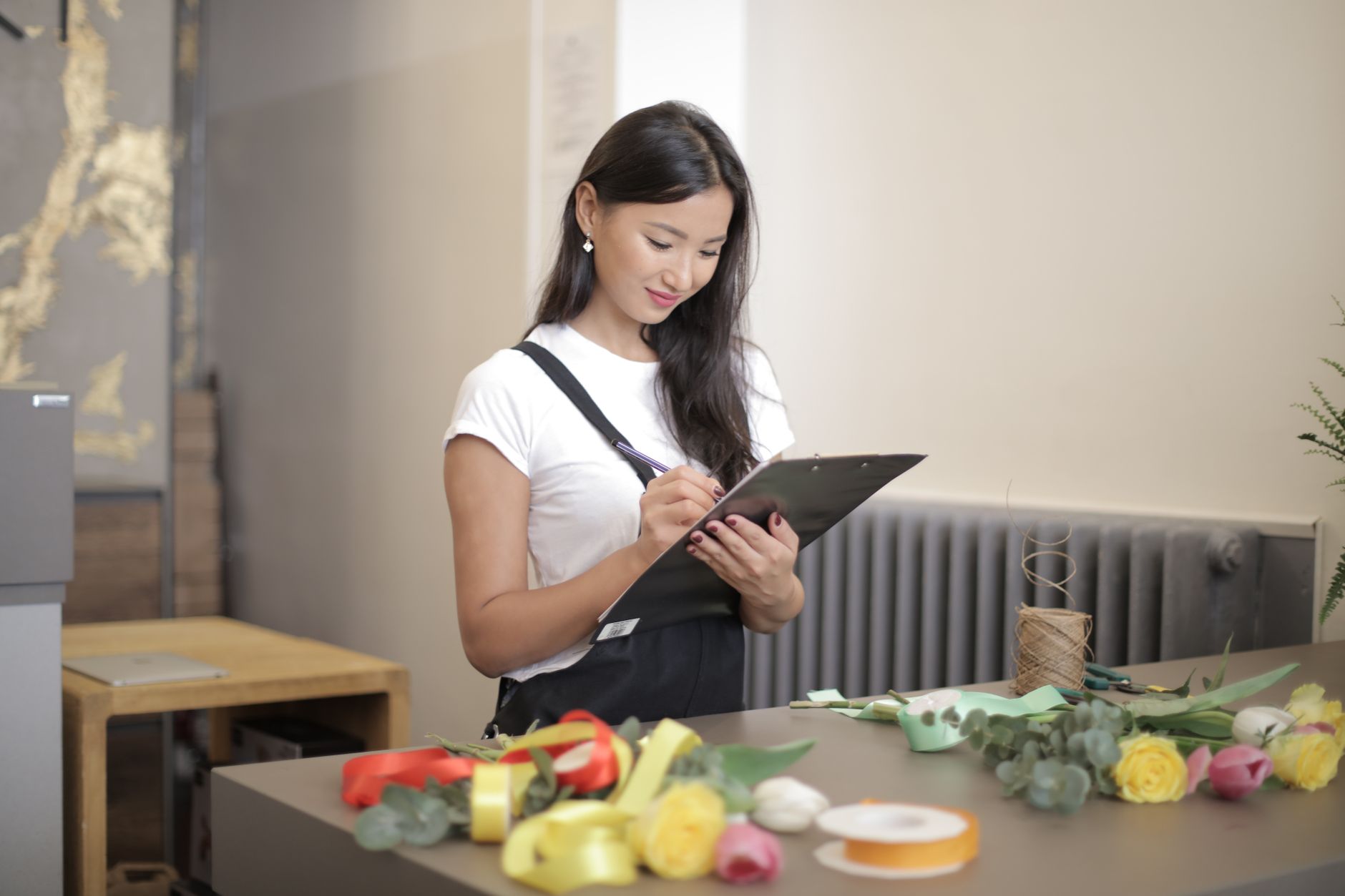 Asian female florist checking inventory in shop with flowers and ribbons on table.