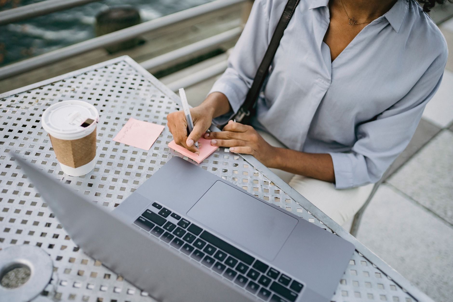 Businesswoman writes notes at outdoor table with laptop and coffee, multitasking efficiently.